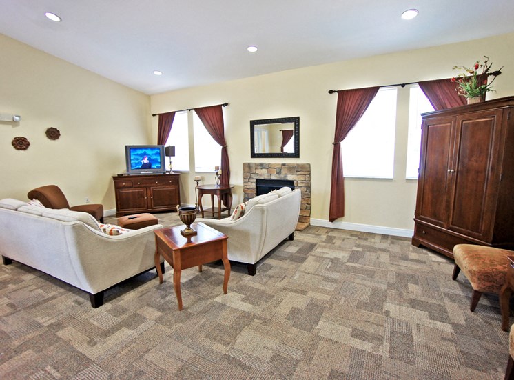 Interior of the clubhouse with carpeted flooring, beige couches, brown chest and wardrobe and end table, tv, and fireplace